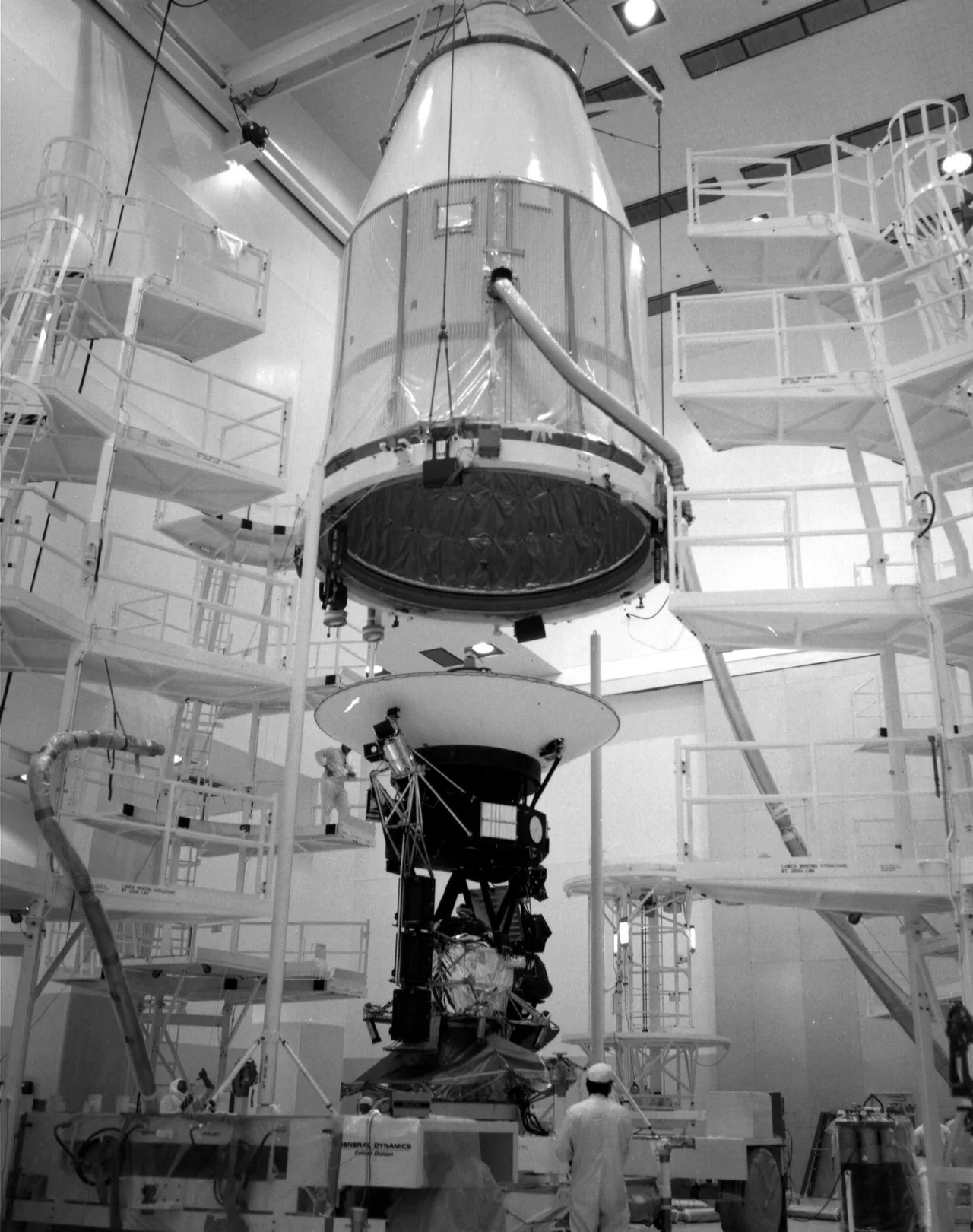 Voyager 2 stands below a raised conical flight shroud in an assembly building, with its large dish antenna and instrument structure visible.