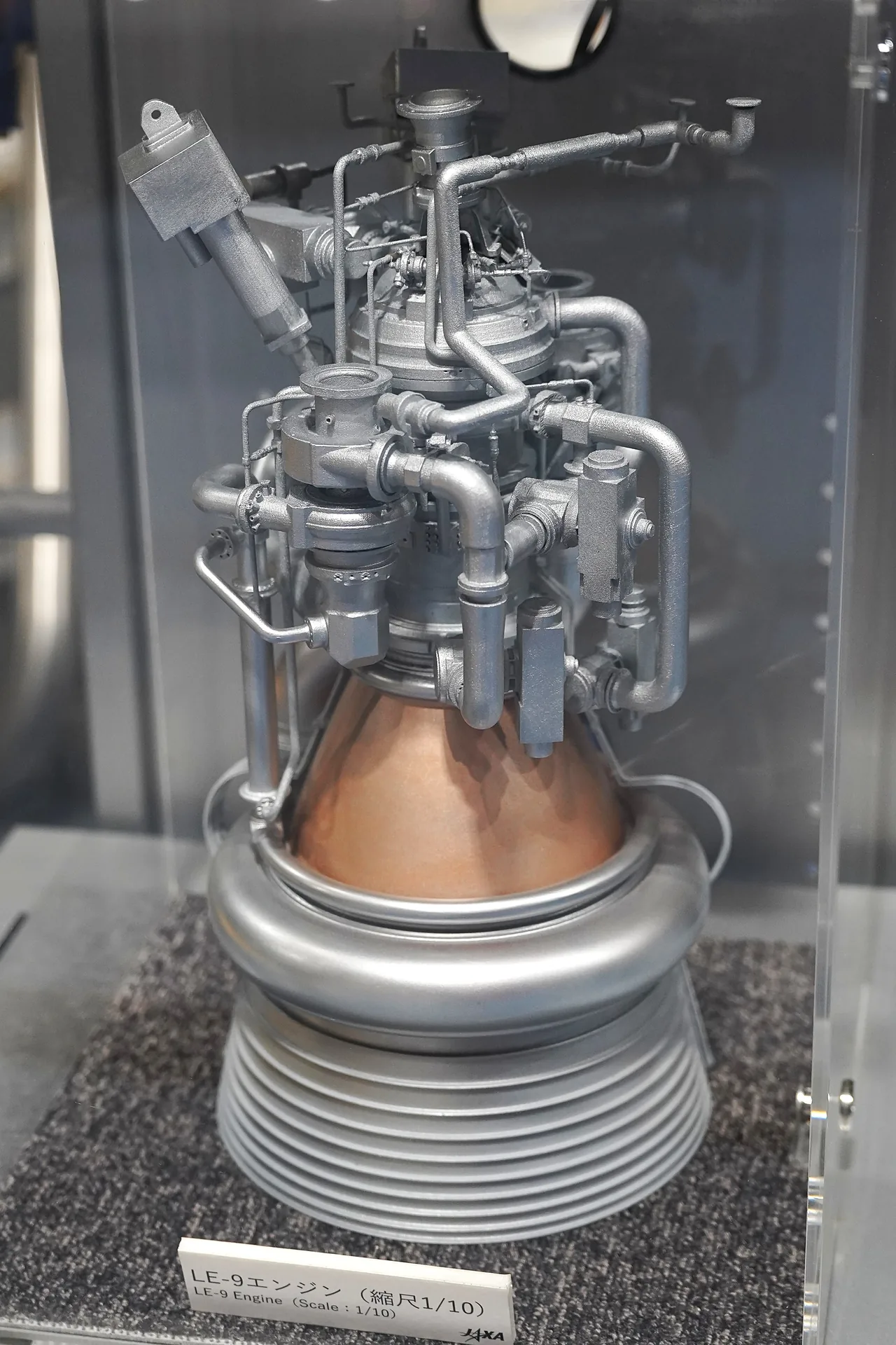 A silver and copper-colored LE-9 scale model stands behind glass, with visible pipes, a nozzle and a sign identifying its one-tenth scale.