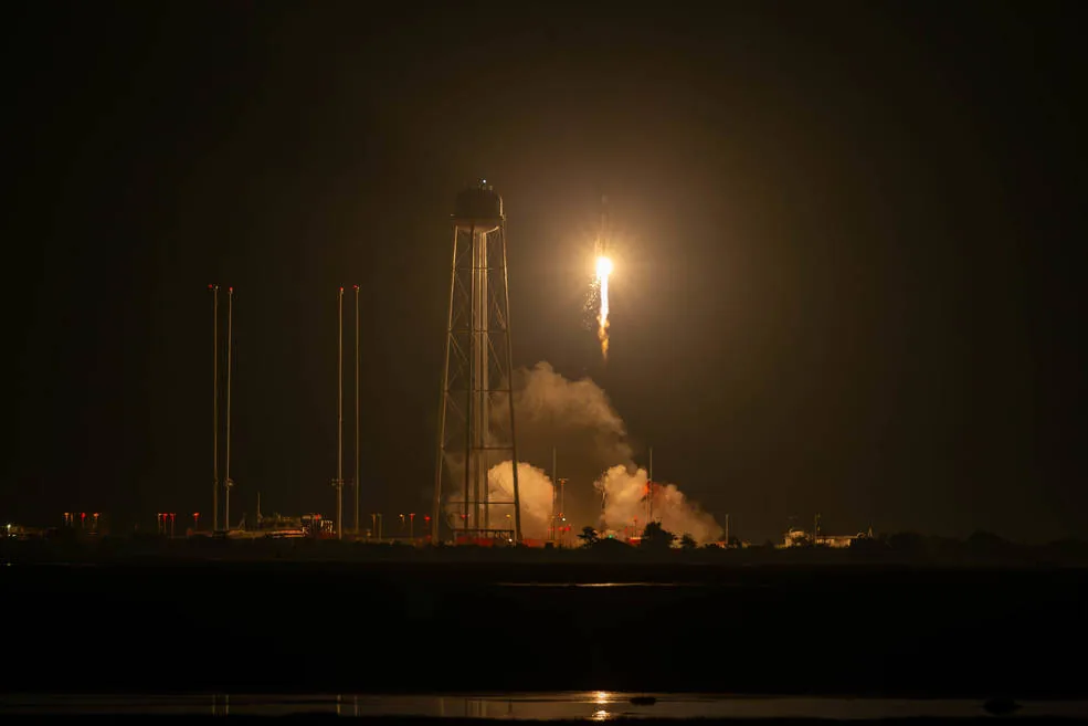 A bright rocket flame rises into the night sky beside a water tower, with illuminated exhaust clouds over the launch pad