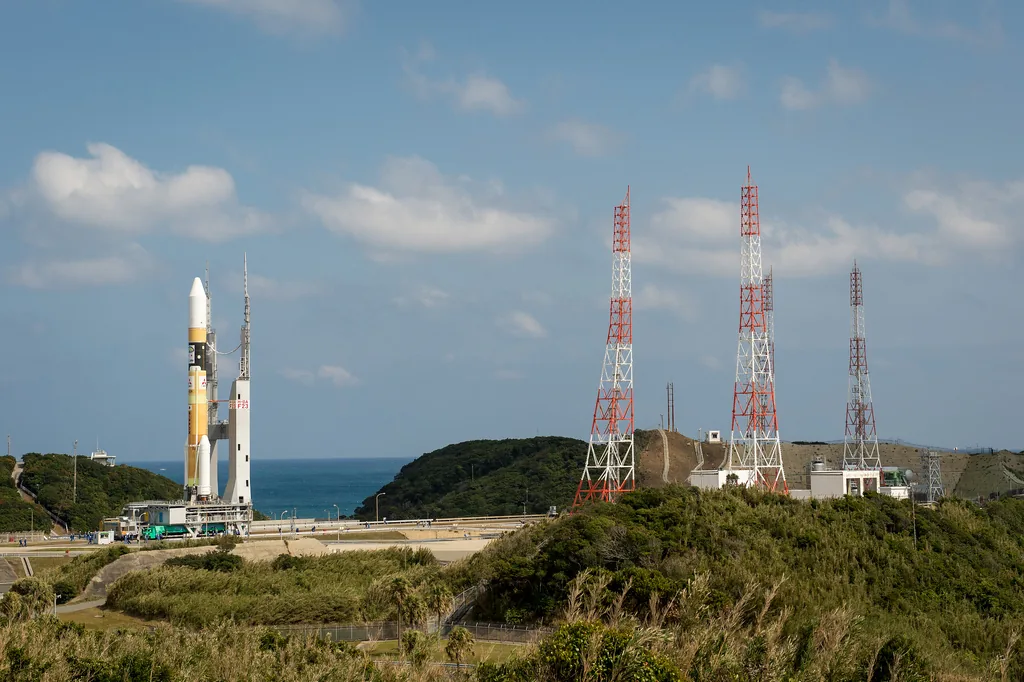 Orange-and-white H-IIA stands on a mobile launcher near the coast, with launch-pad towers across the road and small ground vehicles below.