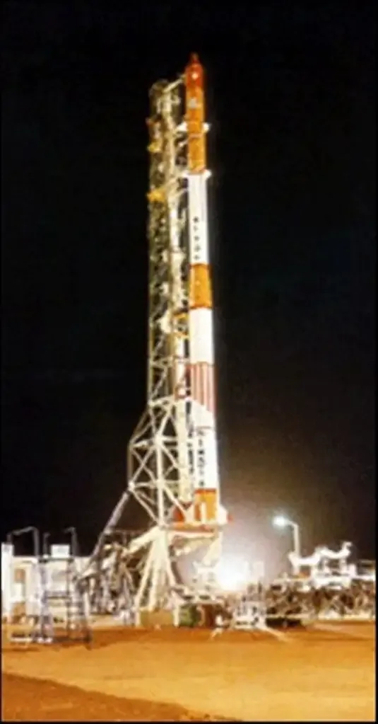 An orange and white four-stage rocket stands beside a lattice service structure at a floodlit launch pad.