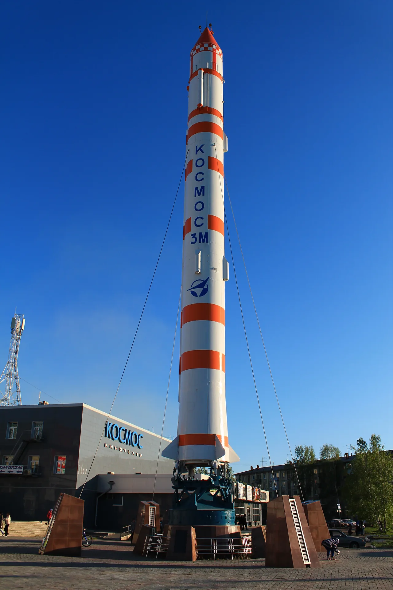 Tall white-and-red Kosmos-3M launch vehicle displayed vertically in an Omsk square beneath a blue sky.