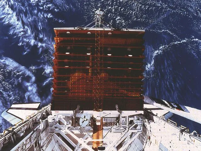 A rectangular solar-array panel extends above a space shuttle cargo bay with clouds and ocean below
