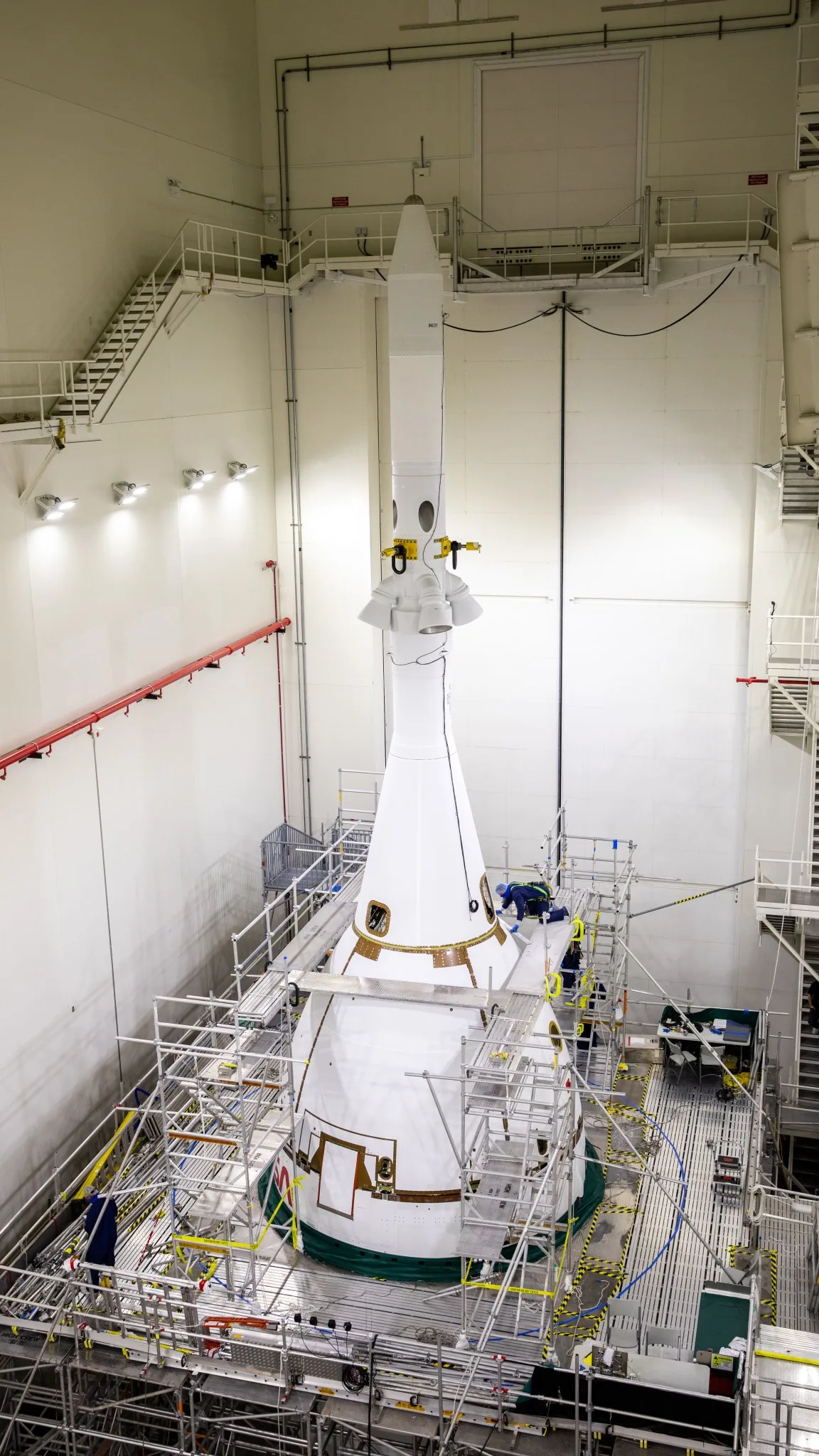 A white launch-abort tower and protective cone stand inside a tall integration building with workers on surrounding platforms