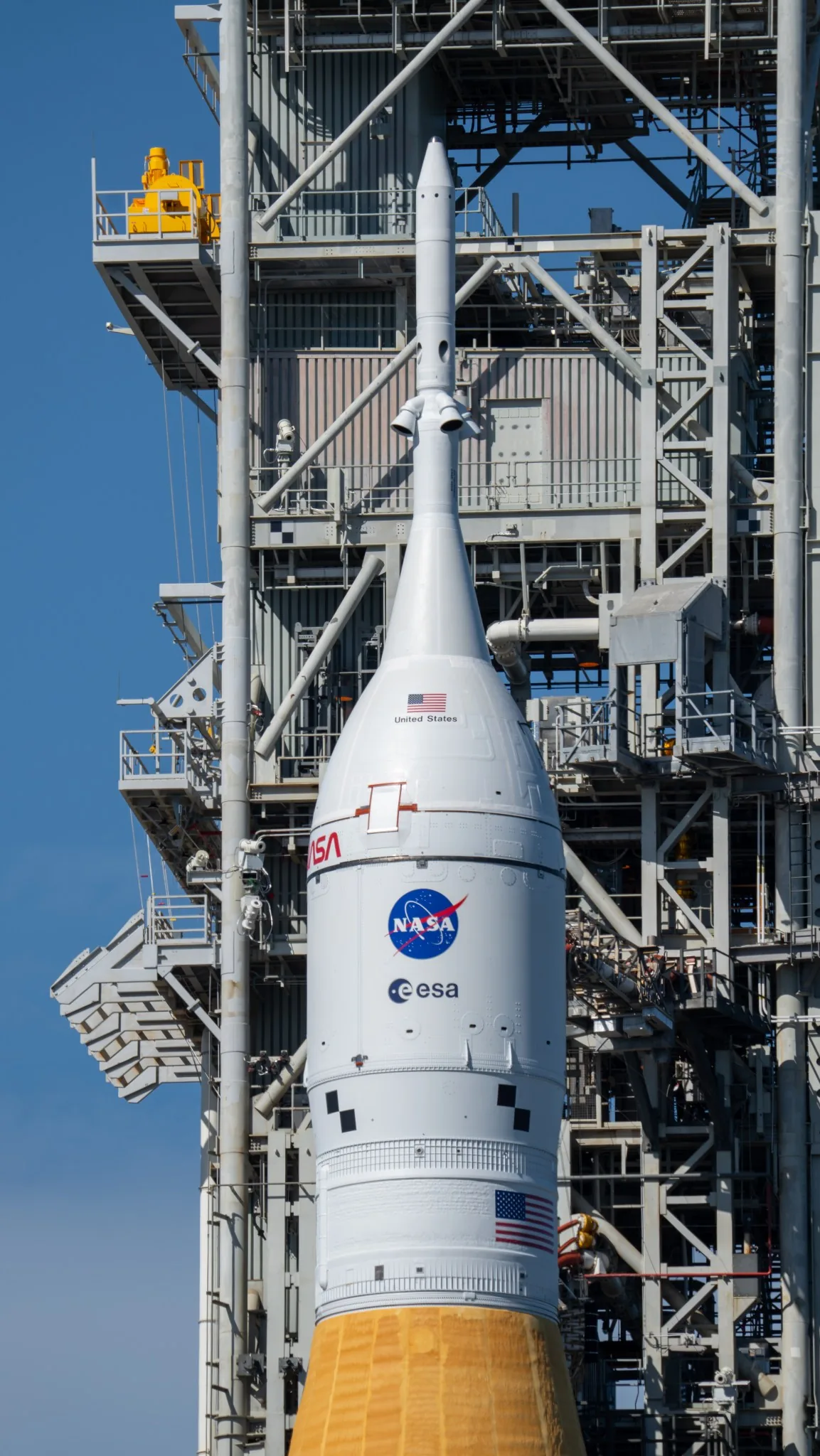 Orion’s white protective cone and launch-abort tower stand beside a steel service structure under a blue sky
