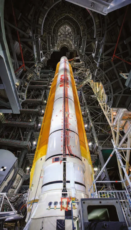 An upward view inside the assembly building shows a white SLS booster beside the orange core stage