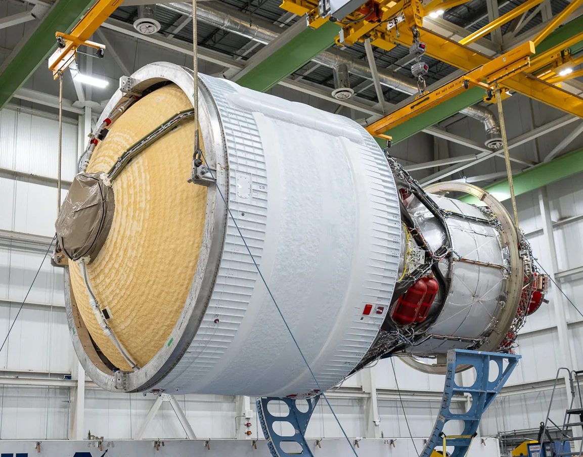 A cylindrical upper stage rests horizontally on blue supports below yellow overhead lifting equipment