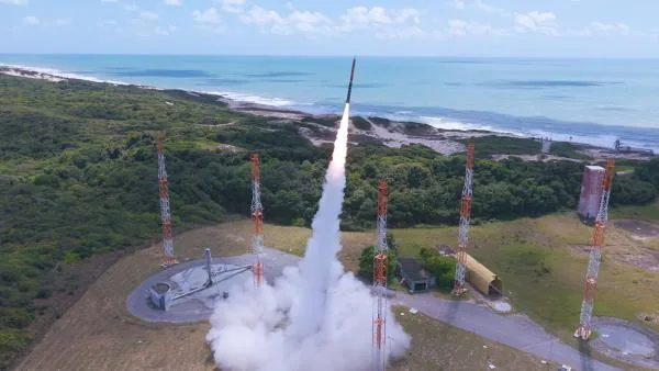 A slender rocket rises on a bright exhaust plume above a coastal launch pad, surrounded by towers, green vegetation and the blue ocean
