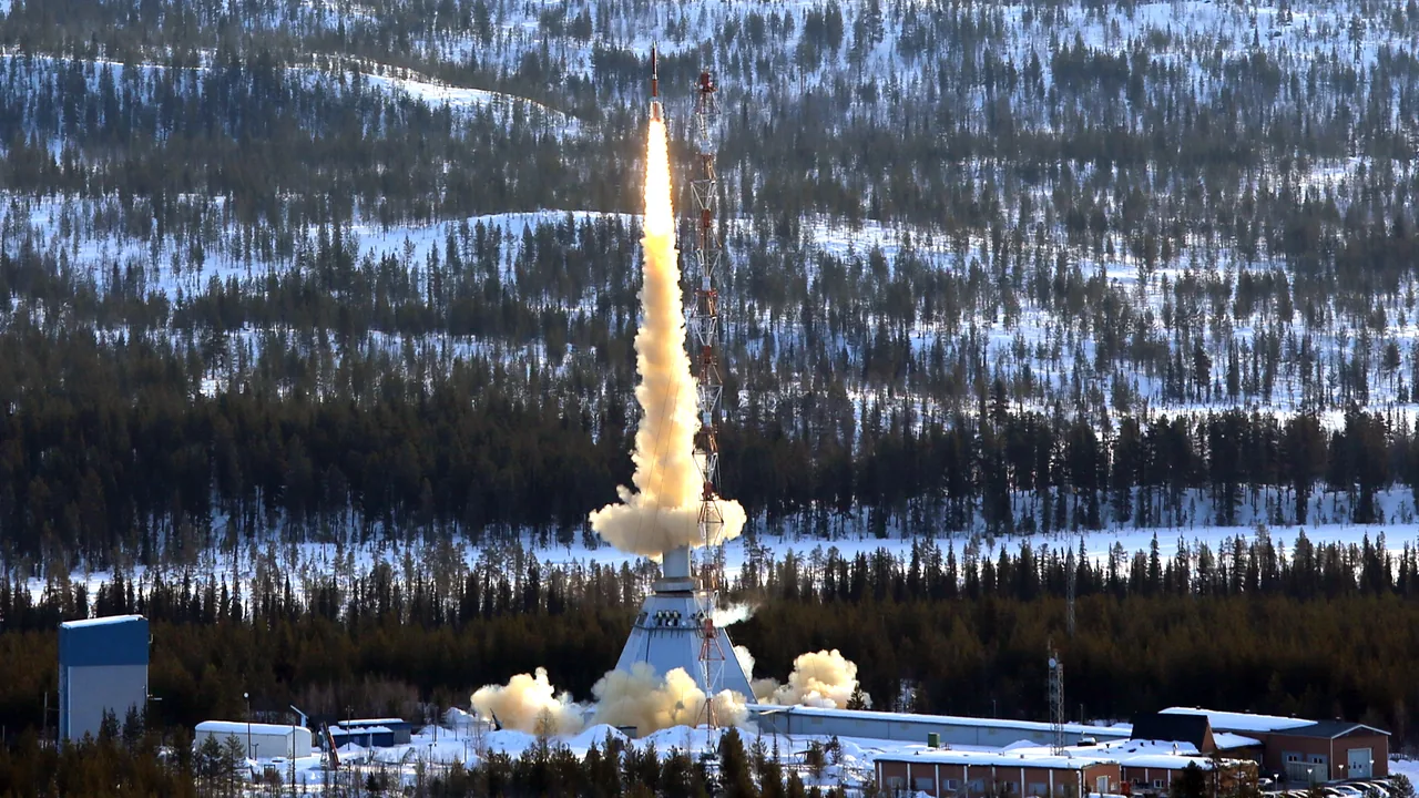 A slender sounding rocket rises on a bright flame above launch buildings in a snow-covered forest